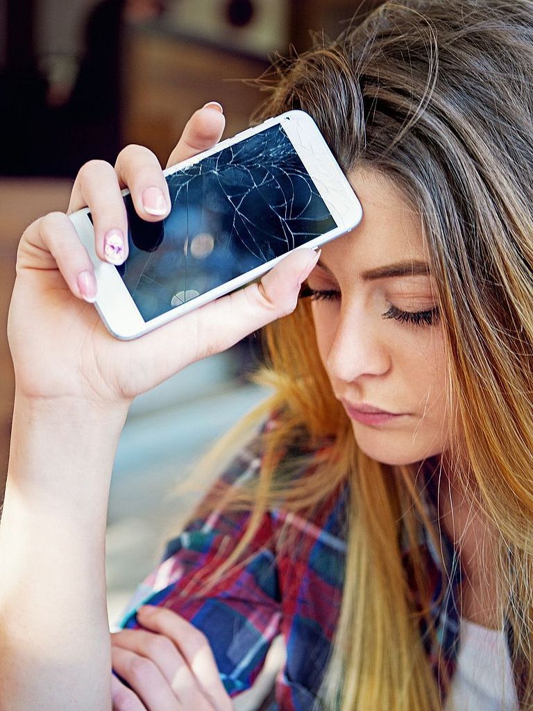 Junge Frau mit traurigem Blick. Sie hält ihr Smartphone mit gebrochenem Display in der Hand