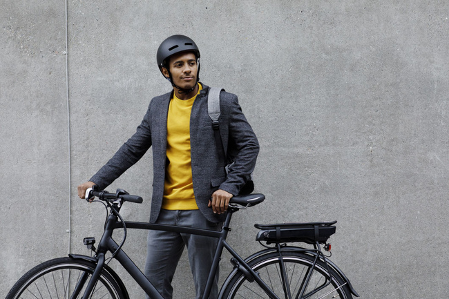 Ein glücklicher Mann steht mit seinem E-Bike vor eine grauen Wand.
