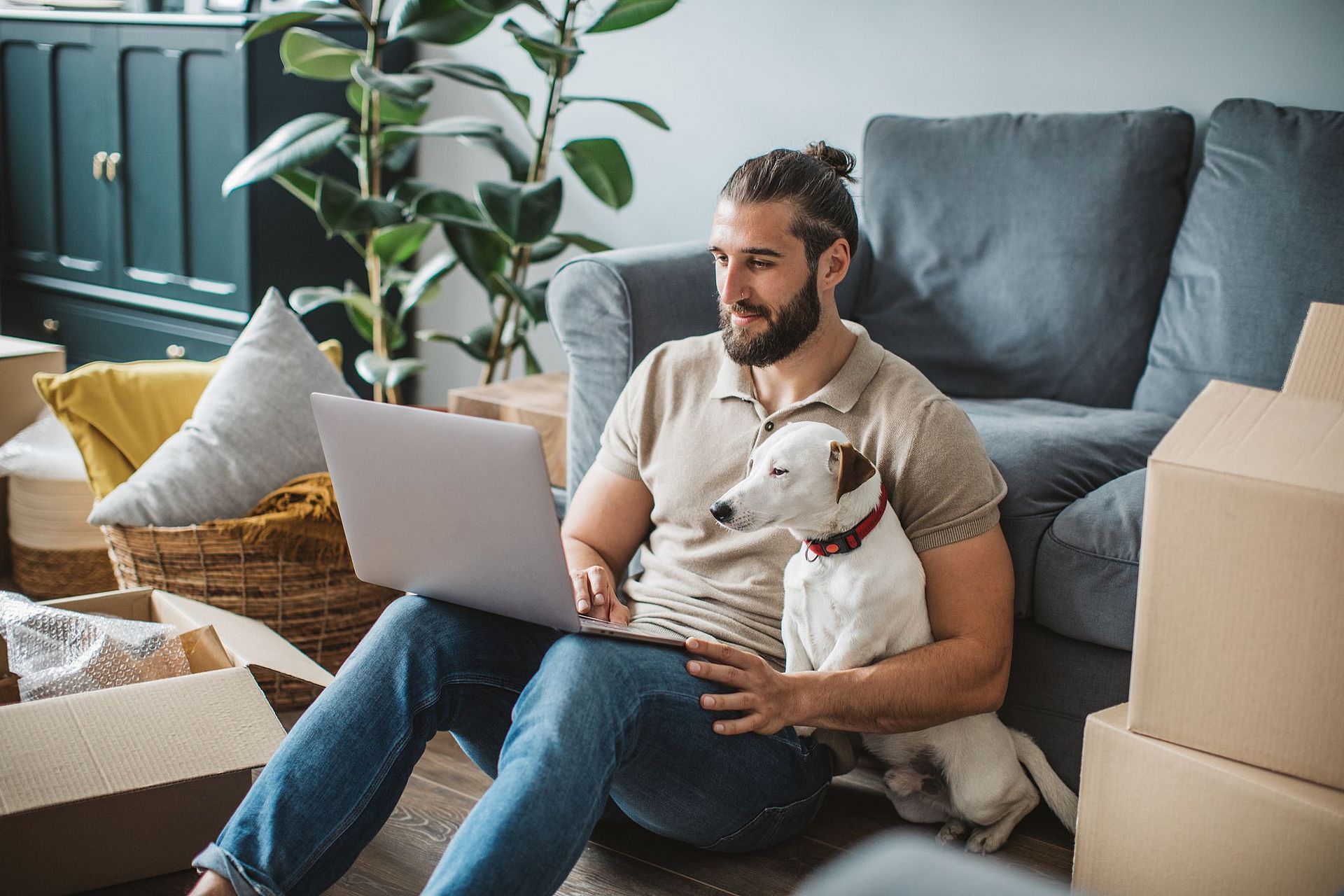 Ein Mann sitzt auf dem Boden vor seiner Couch: in einem Arm sein Laptop, im anderen seinen Hund