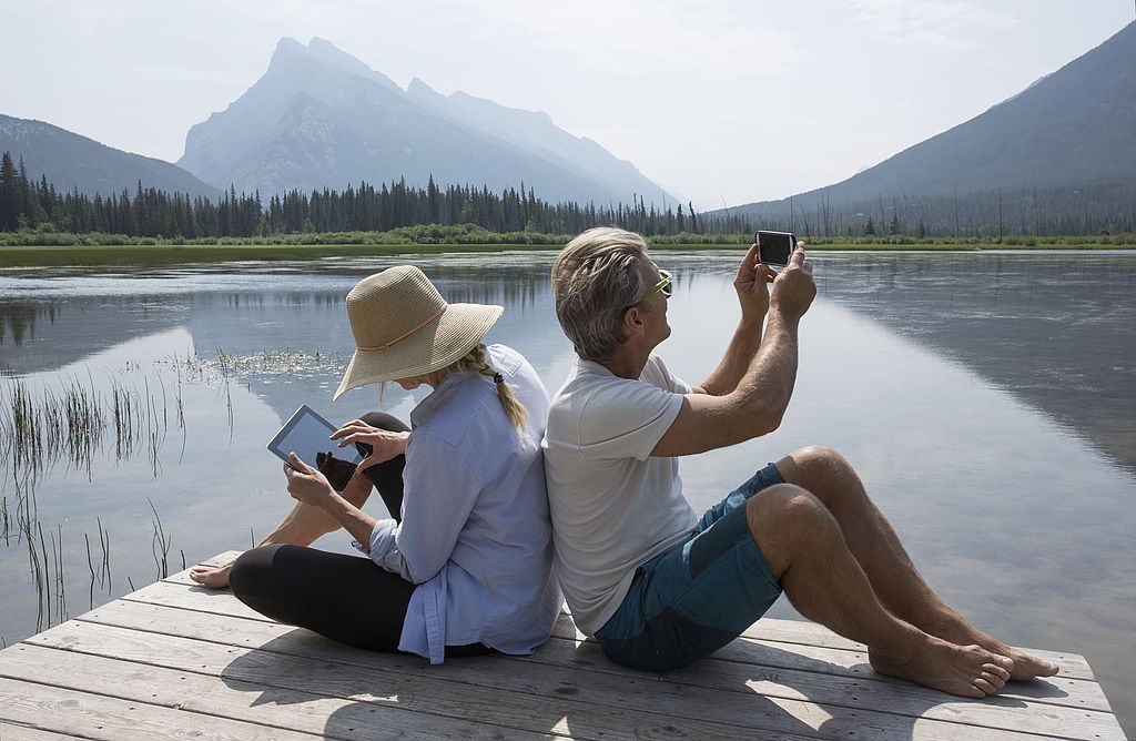 Ein Paar auf einem Steg. Sie liest auf dem Tablet. Er fotografiert mit dem Smartphone